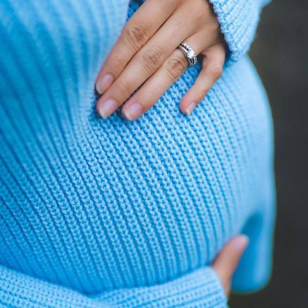 Eine Frau in hellblauem Pullover zeigt stolz ihren Schwangerschaftsbauch